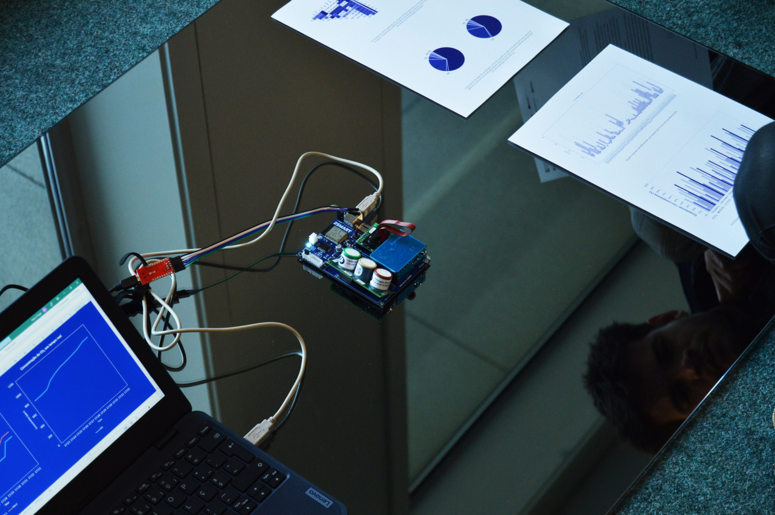 A photograph of the ‘HMV_II’ exhibition. The image shows a computer connected to an Arduino displaying white line graphs on a blue background. The computer is positioned on the floor on top of a mirror, which reflects a viewer’s face.