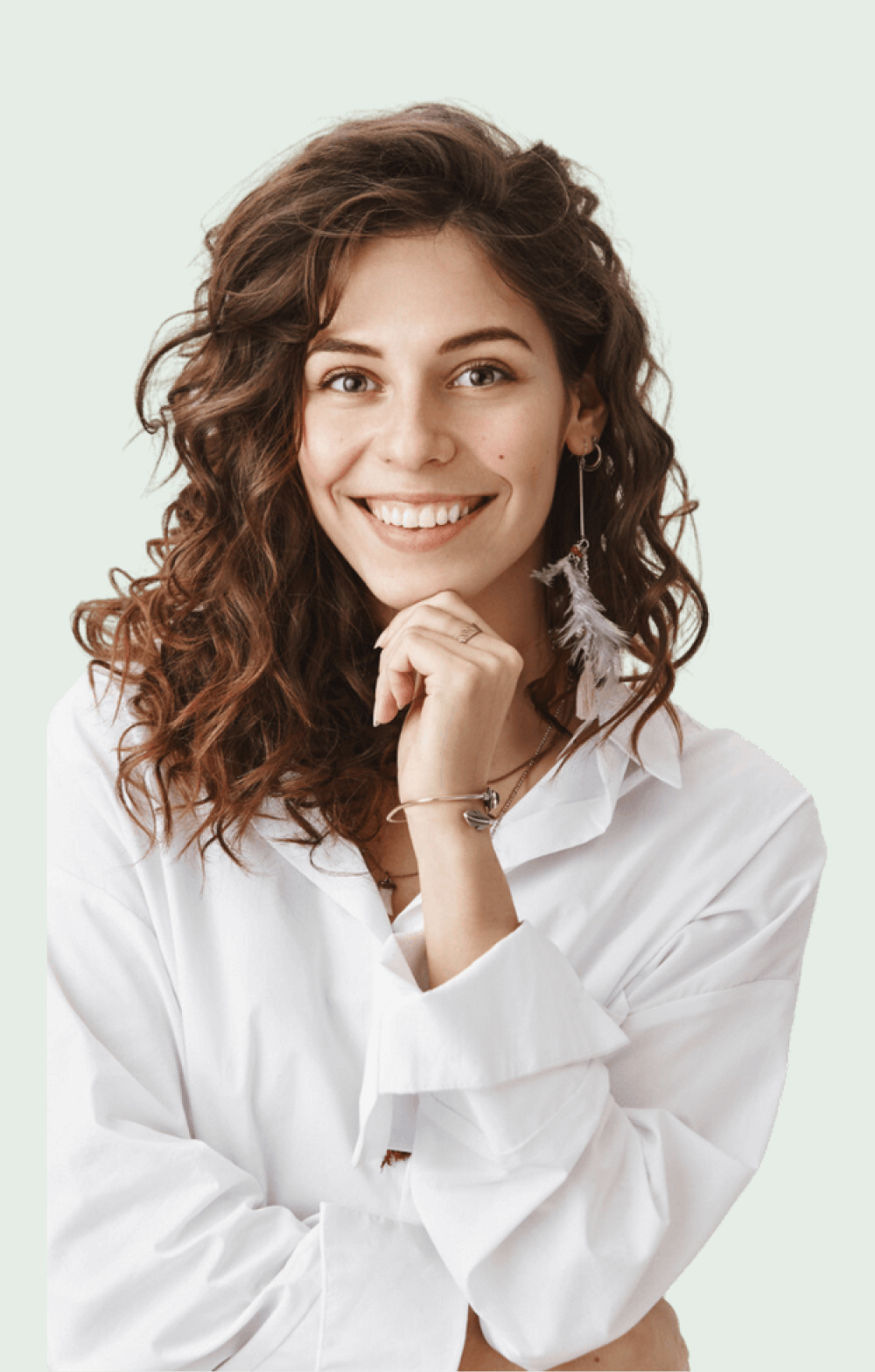 A young woman with curly brown hair and a bright smile is pictured. She is wearing a loose white shirt and a feather earring. She rests her chin on her hand, conveying a cheerful demeanor.