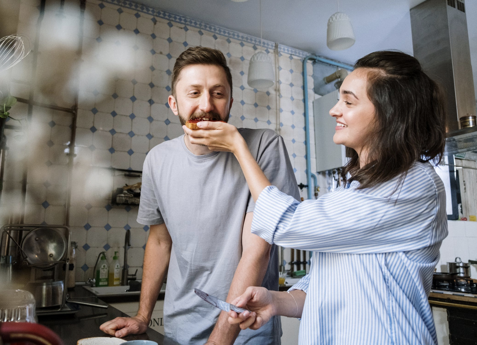 A smiling woman feeds a man a piece of toast in a kitchen, capturing a moment of intimacy and affection.