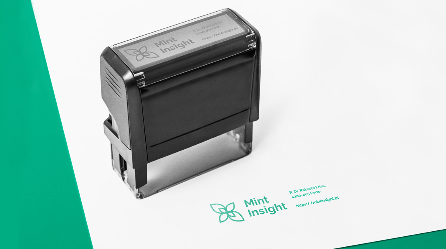 A black self-inking stamp with the Mint Insight logo and contact information on a white piece of paper. The paper is resting on a green surface